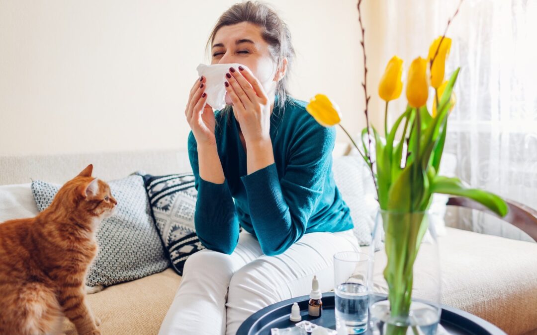 A woman sits indoors sneezing into a tissue and surrounding by allergens such as a cat and flowers.