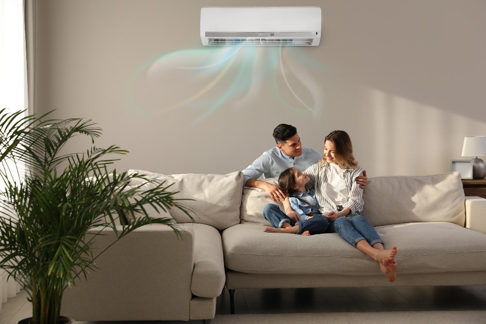 A happy family sits on a couch resting comfortably under an air conditioning unit.